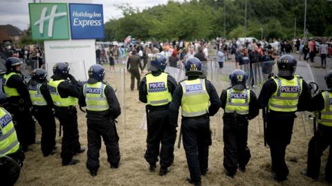 A line of police officers with riot shields and wearing helmets, with members of the public in the background near a Holiday Inn Express hotel. 