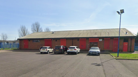 Thornaby Community Centre is a long single-storey brick-building with red doors and shutters pulled down over the windows. Four cars are parked in front of it.
