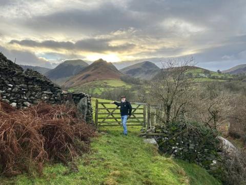 A man wearing a cap and with a white beard and wearing a black top and blue jeans stands in front of a gate with his right hand resting on the top of it. The gate is in the middle of a stone wall and there are fields and hills behind him