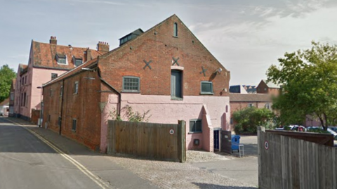 Front of a long building that is made of red bricks and painted pink at the front. There are two fences leading to a car park with trees. 