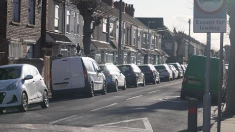 A residential street lined with terraced houses and leafless trees. Cars and vans are parked tightly along both sides of the road. A road sign is visible in the foreground on the right.