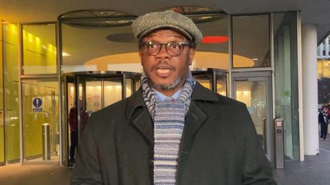 Charles Ehikioya is seen wearing a flat cap, glasses, a striped scarf, and a dark coat is speaking while holding a piece of paper in front of a modern building while delivering a statement. 