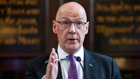 John Swinney, who is wearing a navy blazer, a purple tie and a white shirt, addressing a press conference. Behind him, blurred, is a board with calligraphy writing on it.