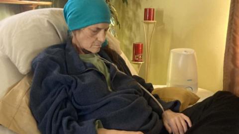 Kelly lies on a hospital bed propped up by pillows and looking down, as if asleep. She wears a blue headscarf on and has a clear tube feeding into her nose. Her hands rest of her lap. 