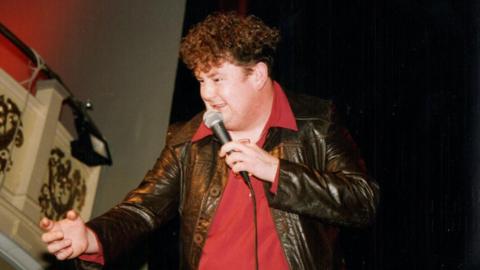 Johnny Vegas wearing a red collared top and leather jacket