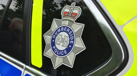 A close-up of a rear window of a police patrol car. The tinted glass is partially covered with a bright yellow sticker and a large Humberside Police logo.