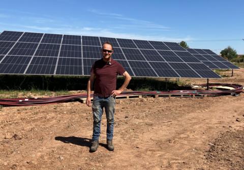 A man wearing a maroon top and jeans stands in front of large solar panels at a farm.