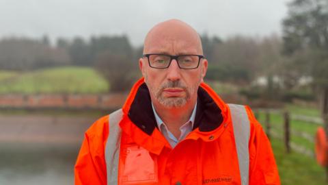 A man in a high-vis orange jacket that says South East Water on it. A body of water and some trees can be seen in the blurry background. He has a stern expression.