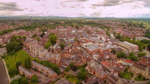 An aerial view of Shrewsbury town centre. There are hundreds of buildings in clusters and a road running through the middle. It is surrounded by patches of green space and trees, with a section of river on the left.