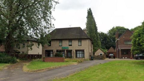 A cream and brown brick building with windows. A short brick wall sits in front, as well as patches of grass. There is a green sign hanging on the front of the building with the text 'The Langleys'.