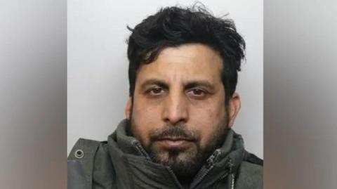 A mugshot of Shahid Hussain who has dark hair, brown eyes and a beard. He appears to be wearing a thick coat.