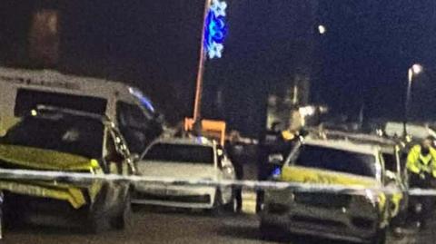 Several police cars and officers on a street in the dark, behind police tape.
