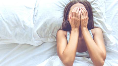 A young woman lies in bed trying to get to sleep. She covers her face with her hands. 