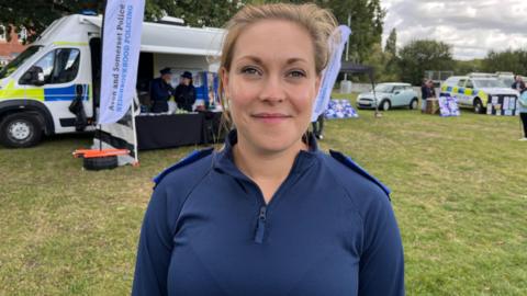 PCSO Bea Gane is looking at the camera, she is smiling and wearing a blue three-quarter zipped jacket with police epiliets on her shoulder. Behind her is a police van with flags flying outside it that say Avon and Somerset Police, Neighbourhood Policing. 
