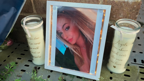 A photo of a young woman inside a white photo frame which has small lights in it. It is propped up against a wall and flanked by two identical white candles.