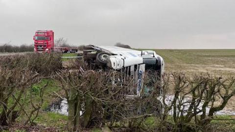 A white lorry on its side in a wet field by the side of the A15. Bare bushes stand in front of it