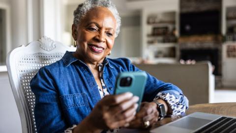 A woman with greying hair and a blue shirt sits at a table, using a phone with a laptop in front of her