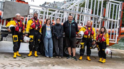 Rachel, Evie and Paul Toms with Looe RNLI volunteer crew members Victoria, David, Clive and Amber. They are stood in front of one of the crew's boats after it has returned from an incident