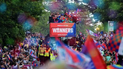 Crystal Palace players can be seen aboard the top deck of an open top bus, surrounded by crowds of fans, many waving red and blue flags 
