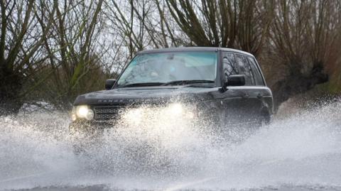A large 4X4 car drives along a flooded road. Surface water is being catapulted into the air as the car drives through it. The Range Rover is a dark colour and has its headlights turned out. Trees are in the background.