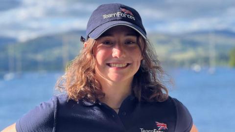 Zara Lachlan wearing a dark blue baseball cap and dark blue polo shirt, both with Team Forces and the Union flag logos. She has shoulder length brown, curling hair and is smiling broadly. Behind her is a blurred out blue sea and hills beyond.