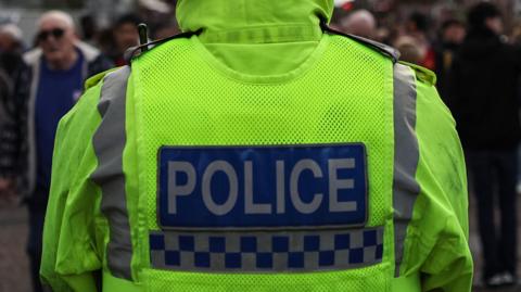 A generic shot of a police officer taken from behind. He is wearing a hi-viz jacket with the word 'police' printed on the back.