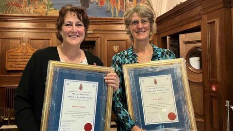 Two award recipients - a woman with brown hair and a woman with blonde hair - standing indoors holding framed certificates with blue backgrounds and gold borders. The certificates feature printed text and a red wax seal. Behind them is a wooden-panelled wall with a shield emblem and a large mural above.