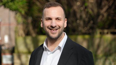 Zack Polanski smiling on a sunny day withy trees and a fence blurred in the background. He is wearing a black jacket and white shirt open at the neck.