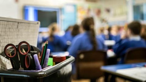 A stock image of children sitting in a classroom. In the foreground there is a container with scissors, pencils and a ruler. The background is blurred and shows the seated children wearing blue uniforms.