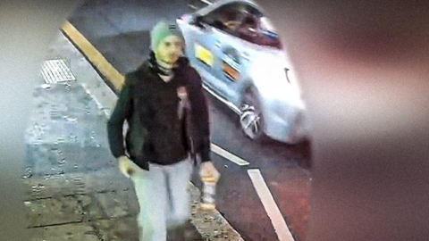 A man wearing blue jeans, a dark jacket and a beanie hat walking down a road in Hastings at night, filmed on grainy CCTV.