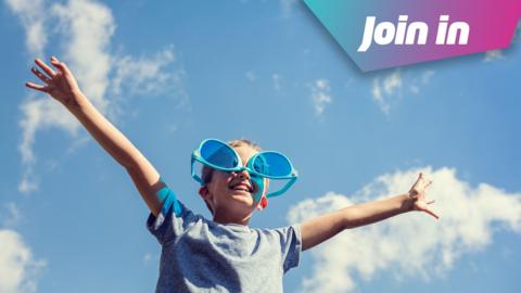Boy wears a blue tshirt and oversized sunglasses upside down on his face. The photograph is taken from an angle below the boy and looking up towards the blue skies with a few white wispy clouds. He holds his arms out, his hands and fingers are spread wide and he smiles