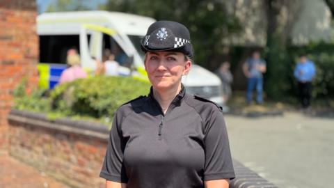 Insp Liz Warner pictured wearing a black police top with a quarter zip and a black police hat. Behind her in the blurred background are people in plain clothes stood next to a police van.
