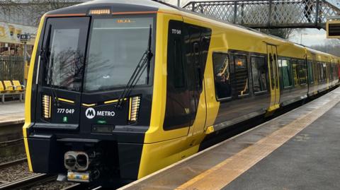 The Merseyrail train is black and yellow, and has stopped at a platform.