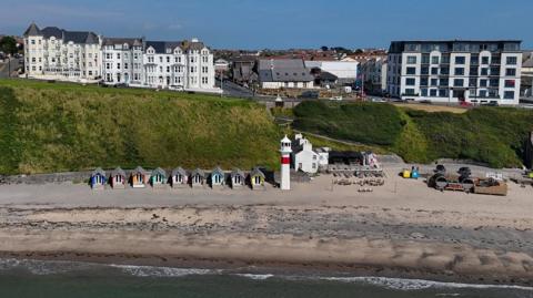 Port Erin beach has buildings on the headland above it and coloured beach huts on the sand.