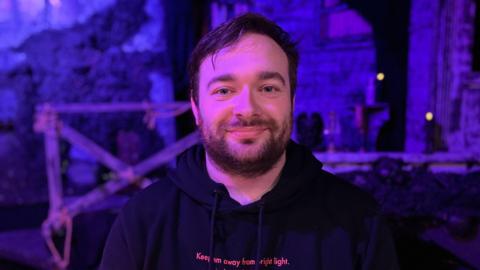 A man with black hair and short beard, he has blue eyes and wearing a black hoodie. He is looking at the camera with a smile on his face.