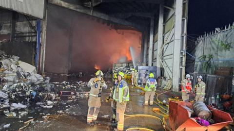 Several firefighters stand in a warehouse building, with flames visible in the background.