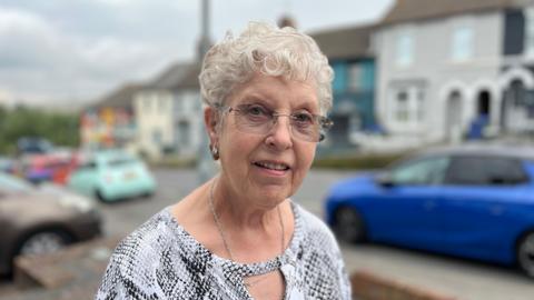 Sylvia Porritt standing next to Lewes Road which has a row of parked cars and terraced houses behind her. She is wearing a silver top and has glasses