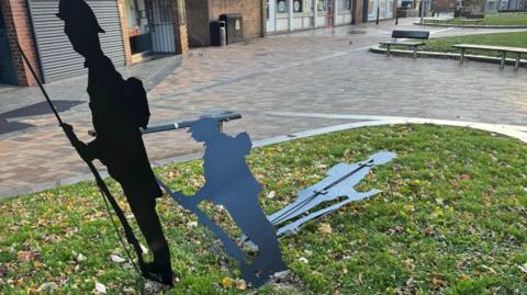 Three black metal silhouette sculptures depicting soldiers. One figure holding a rifle with a bayonet is standing upright while the second figure is bent lower and the third one near the ground. They are placed on a small grassy area with scattered autumn leaves, surrounded by a paved pedestrian space.