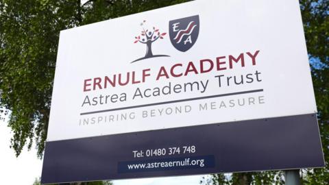 A close-up of a dark blue and white Ernulf Academy sign outside the building against a tree. It says Ernulf Academy, Astrea Academy Trust, inspiring beyond measure and gives the telephone number and website address. 