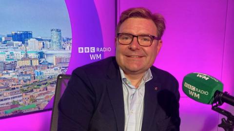 Birmingham City Council leader John Cotton sits in the ˿ Radio WM studio in front of a green microphone and a purple backdrop of the Birmingham city centre skyline