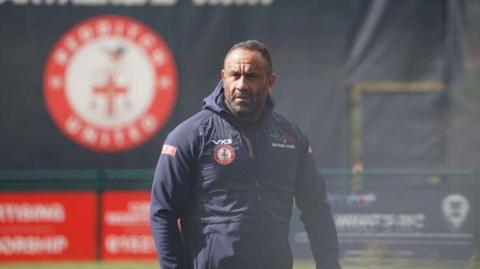 A man wearing a navy, hooded jacket stares off into the distance. The background is out of focus but a blurred Redditch United logo can be made out