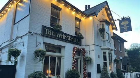 An exterior view of the Wheatsheaf pub in Ewell. It has hanging baskets outside and a bauble display around the door with a black sign and gold lettering.