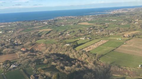 An aerial view of Guernsey from an aeroplane flying in the sky. There is lots of green space and the sea is also visible at the top of the photo.