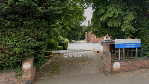 Two brick walls in front of large green trees and bushes flank an entryway with a yellow barrier in front of it. Beyond the barrier is a car park and a large hospital building. A blue and white sign at the entrance reads "Bloxwich Hospital, main entrance"