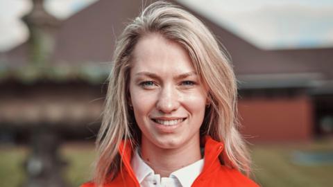 A head and shoulders view of a smiling Lottie Fry, who has blonde, shoulder-length hair and wears a red jacket and white shirt.