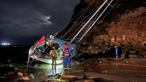 A yacht leaning on its side on a beach with one member of a rescue team on it, and four more on the sand. They are wearing life jackets, helmets and hi-vis clothing. The boat appears to have run aground and strikes a dramatic scene against a darkened sky. 