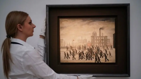 A woman auction worker, wearing a white shirt, holds up the framed LS Lowry Going To The Match rugby league painting