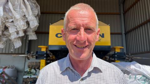 A man in a checked shirt poses and smiles for the camera with yellow farm machinery behind him