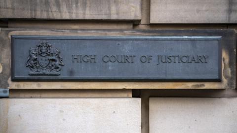 The sign for the High Court of Justiciary outside the High Court in Edinburgh. The sign features black writing on a black background with the UK coat of arms next to it and is attached to a light stone brick wall.