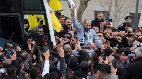 A man carried by a large crowd of people, wearing a grey jumper, a white bus in the background